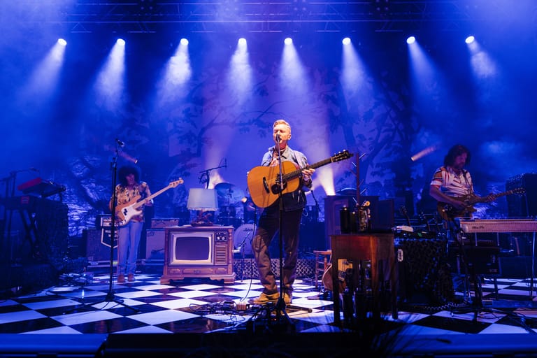 Guitarist performing centre stage with band under blue spotlights on a checkered floor.