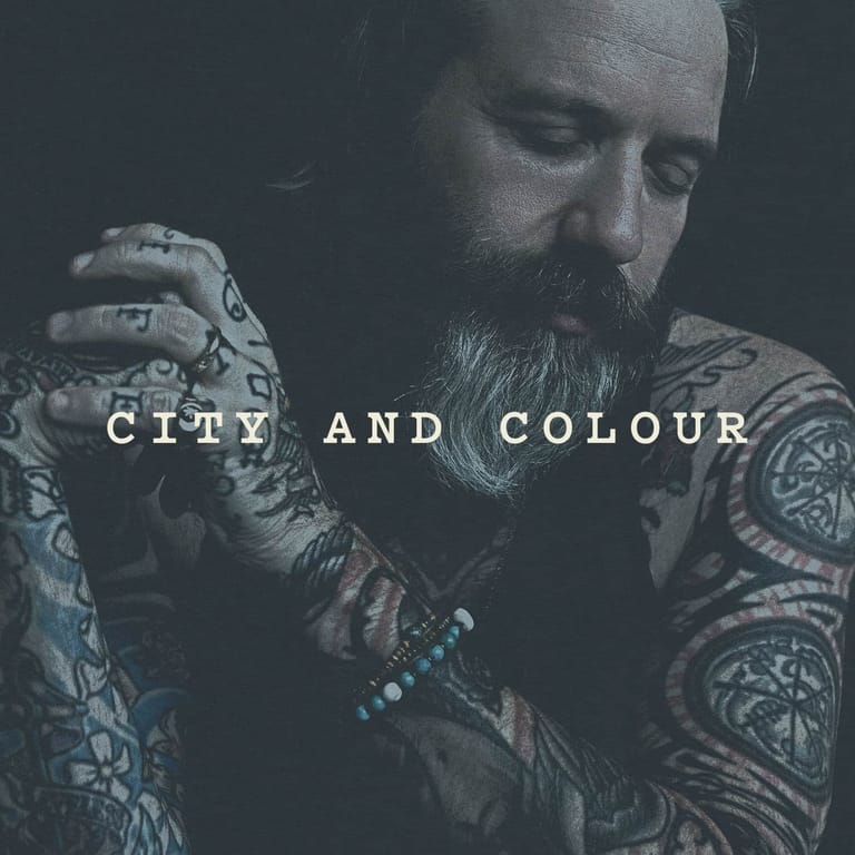 Bearded man with extensive tattoos on both arms displays jewellery against a dark background, with "CITY AND COLOUR" text overlaid.