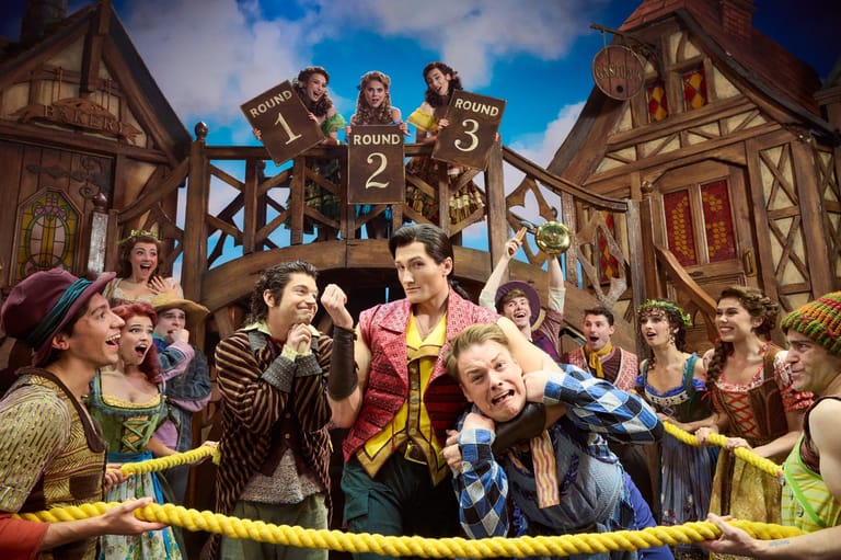 Performers in medieval costumes on a theatrical stage set with timber-framed buildings and a crowd of extras.