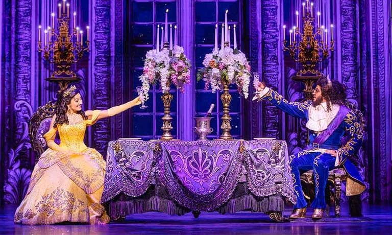 Two performers in formal gowns flank a candelabra on an ornate stage set bathed in purple lighting.