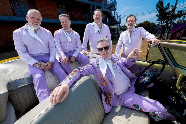 Five people in coordinated purple outfits pose together in a vintage convertible car outdoors.