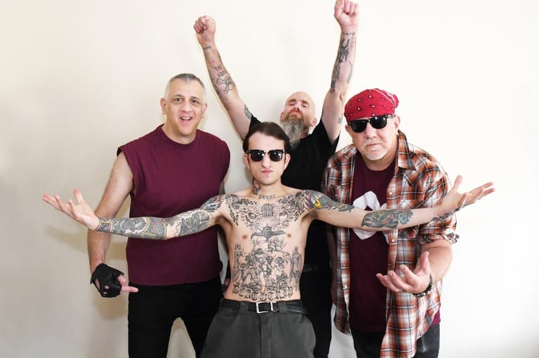 Four band members with tattoos pose enthusiastically against a white background, arms raised and spread wide.