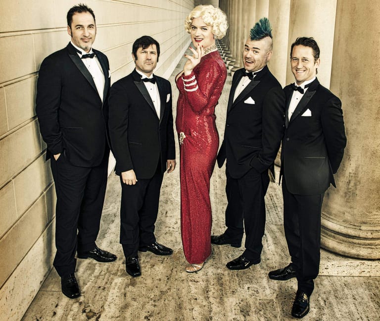 Five performers in formal evening wear stand together in an elegant corridor, the woman in a striking red sequined gown.