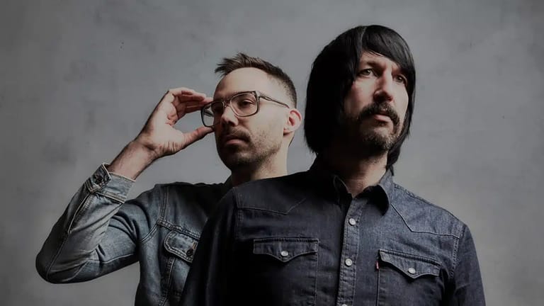 Two men in denim shirts pose against a neutral grey background, one adjusting his glasses.