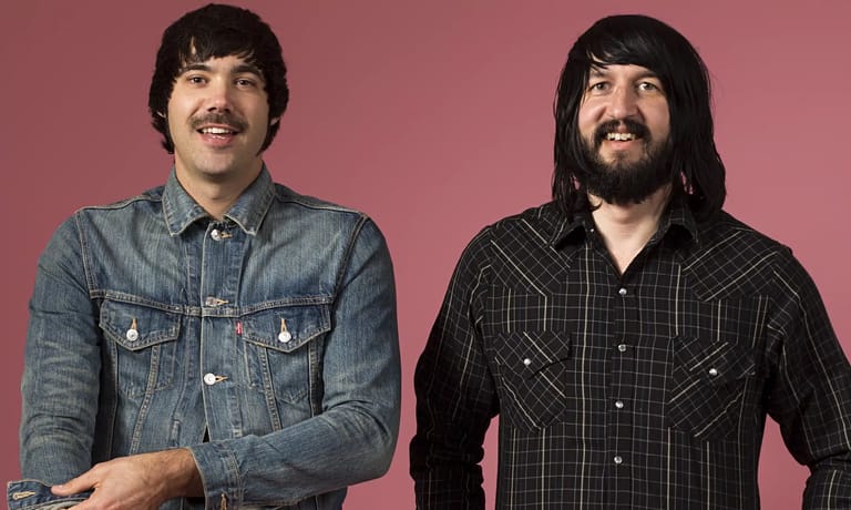 Two men with dark hair and facial hair pose smiling against a pink background, one in denim and one in a black plaid shirt.