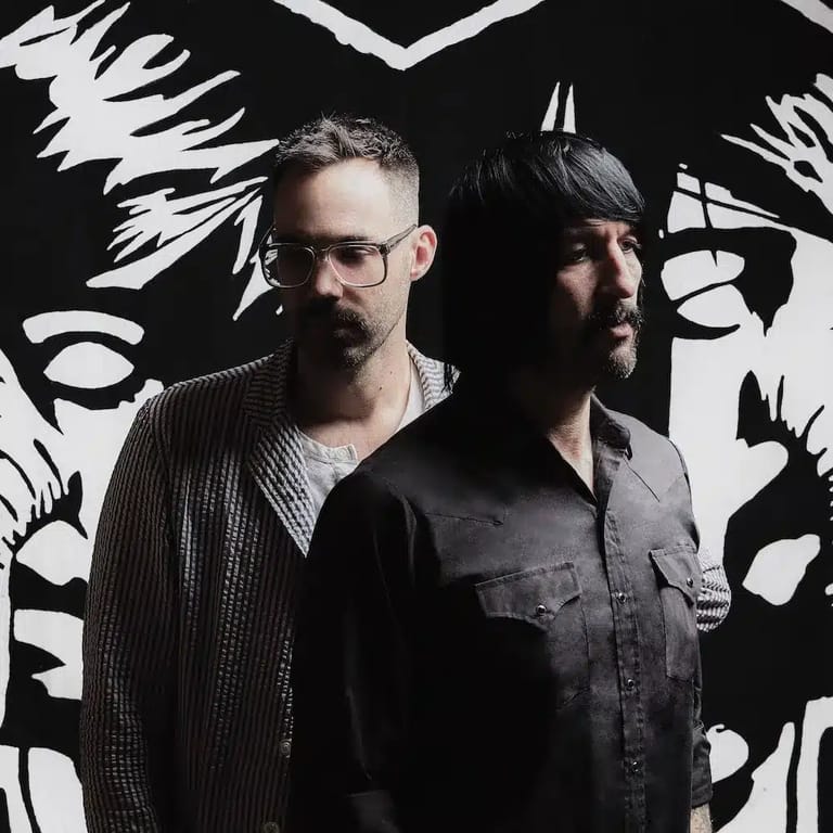 Two men stand against a dramatic black and white graphic backdrop featuring stylised face portraits.