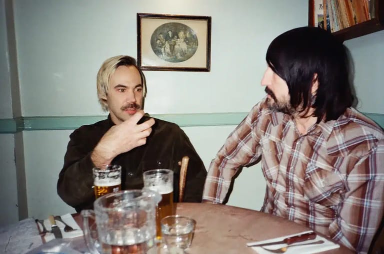 Two men sit at a table with drinks, engaged in conversation in an indoor setting.