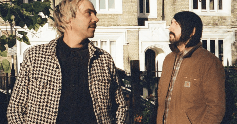 Two men in casual jackets converse outside a stone heritage building.