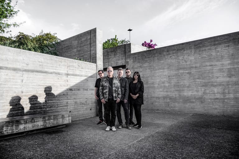 Five people in dark clothing stand in a modernist concrete courtyard beneath overcast sky.