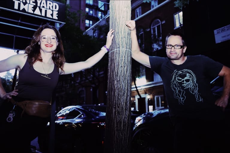 Two people stand on either side of a textured column with arms raised in an urban nighttime setting.
