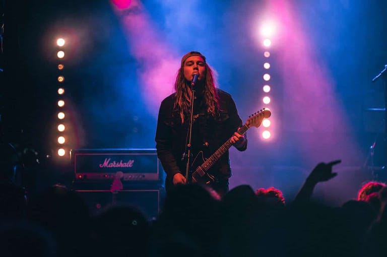 Guitarist performing on stage under pink and blue spotlights with Marshall amplifier visible behind.