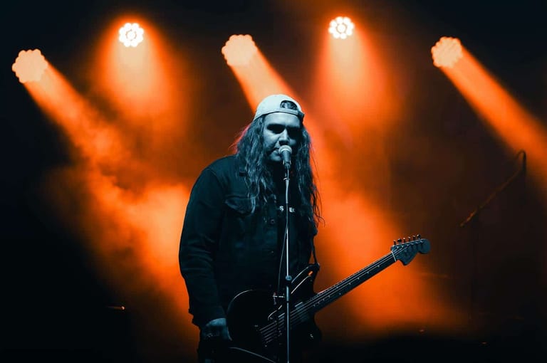 Musician with long hair performs at microphone beneath orange stage lights and beams.