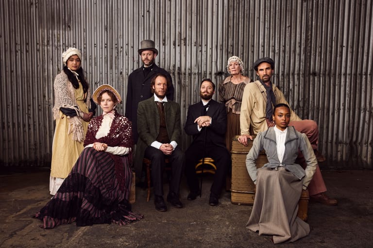 Ensemble of nine performers in period costume posed against corrugated metal backdrop.