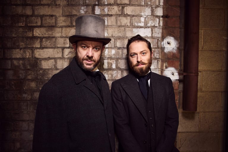Two bearded men in dark suits stand against a brick wall lit by dressing-room mirrors.