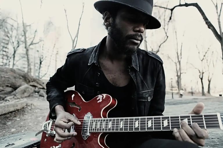 Musician in a black cowboy hat playing a red semi-hollow electric guitar outdoors among bare trees.