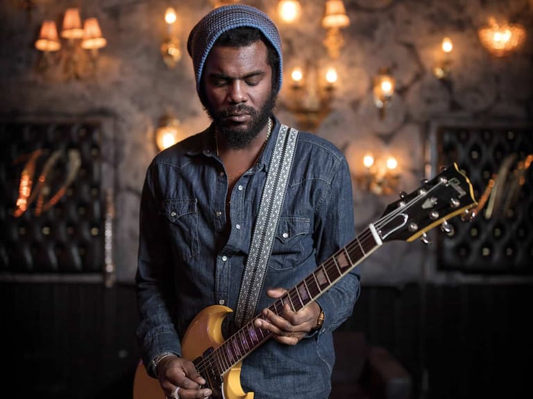 Musician in denim shirt and beanie holding acoustic guitar against warm-lit backdrop with string lights.