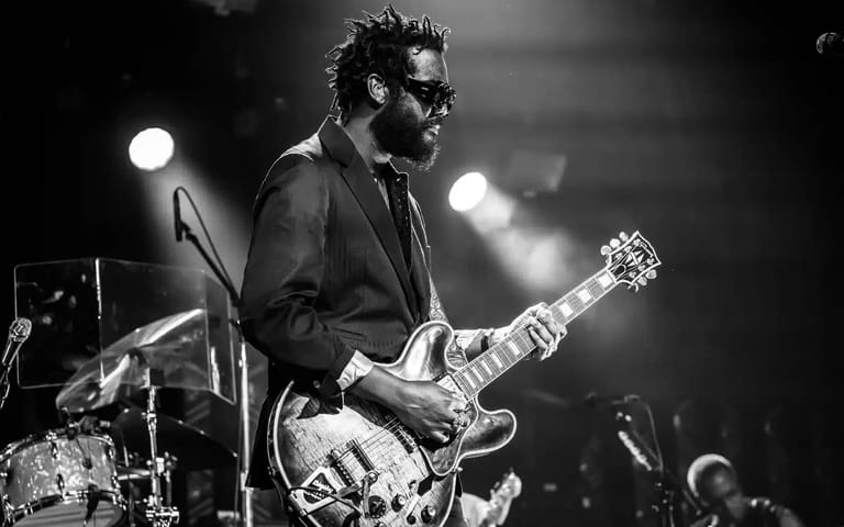 Guitarist in sunglasses and dark suit performing on stage under spotlights during a live concert.