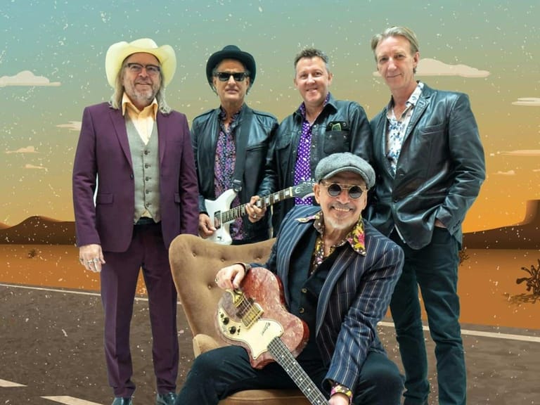 Five musicians in Western-style outfits pose together against a desert sunset backdrop, one seated holding an electric guitar.