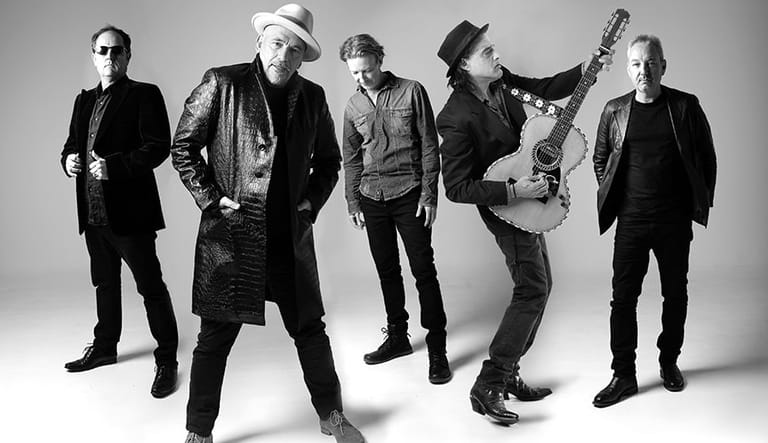 Five band members in dark suits and hats pose together against a grey background, one mid-leap with guitar raised.