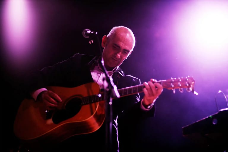 Bald musician playing acoustic guitar under purple stage lighting during a live performance.