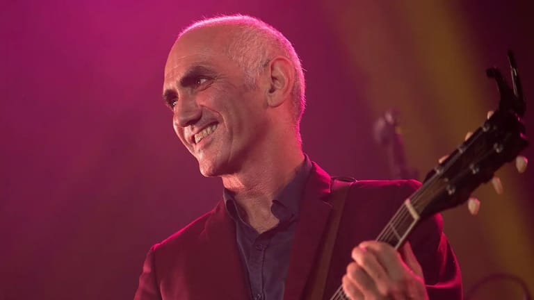 Bald musician with guitar smiling under pink stage lighting during a live performance.