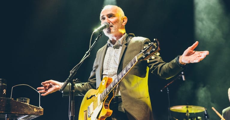 Bald musician playing yellow electric guitar at microphone with arms outstretched on dimly lit stage.