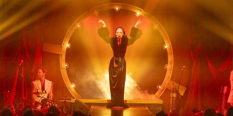 Performer in red silhouetted within glowing circular frame on stage, arms raised in triumph.