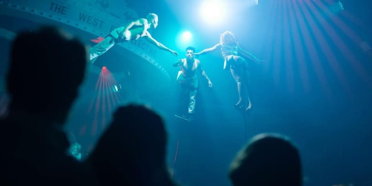 Silhouetted audience members watch performers suspended in mid-air against blue stage lighting.
