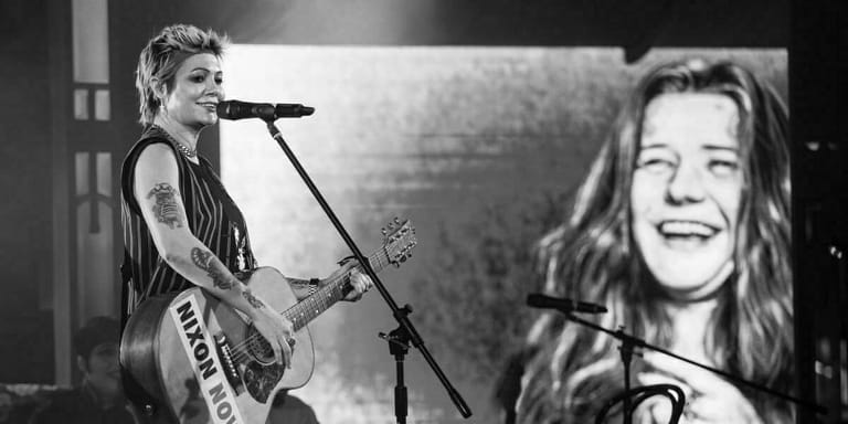 Musician performing with electric guitar at microphone beside large projection screen in black and white.