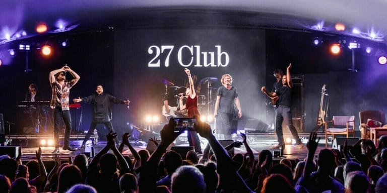 Band performing on illuminated stage at 27 Club venue with audience members raising phones and hands.