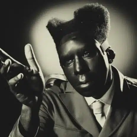 Performer in sharp suit and tie gestures expressively toward camera in dramatic black and white portrait lighting.