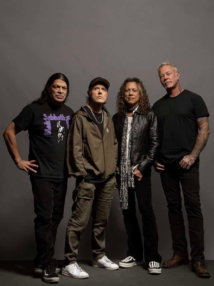 Four musicians in casual black and earth-toned clothing stand together against a neutral grey backdrop.