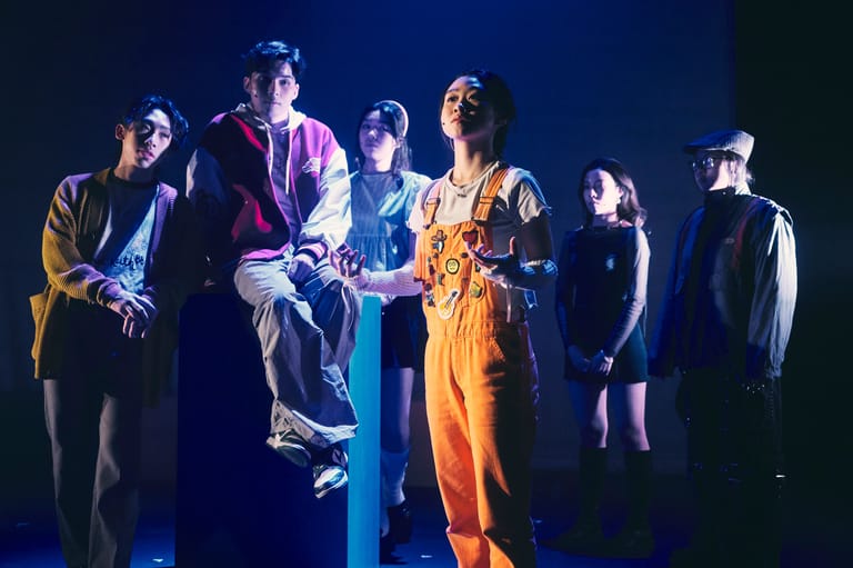 Ensemble of performers in colourful costumes stand together on a dimly lit stage under blue spotlights.