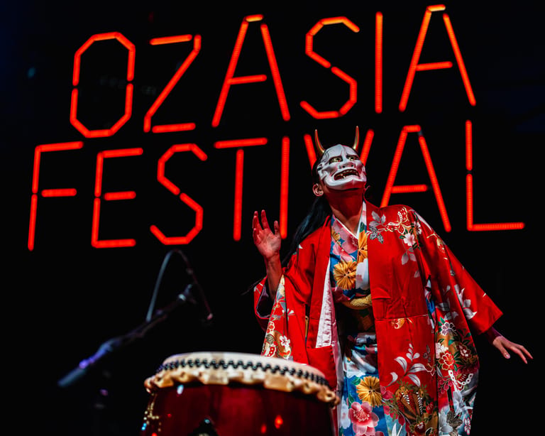 Performer in vibrant red kimono and dramatic makeup gestures expressively before glowing Oz Asia Festival signage.