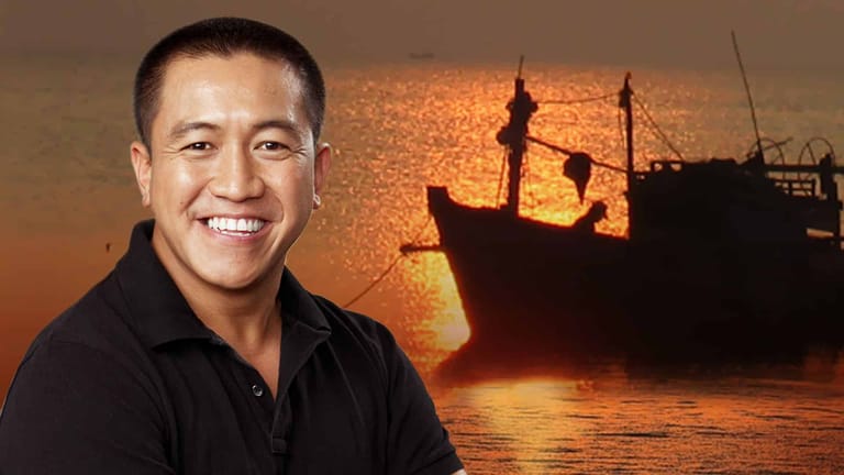 Man in black shirt smiling at camera with silhouetted fishing trawler and golden sunset water behind him.