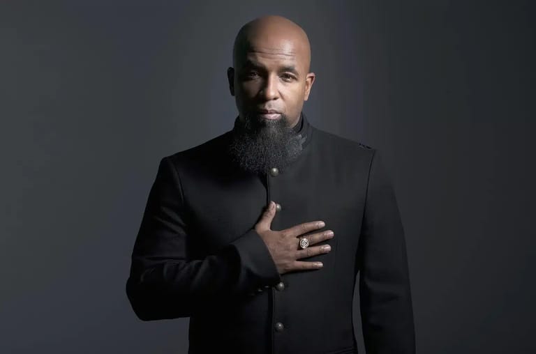 Man in black blazer with hand on chest against neutral grey background.