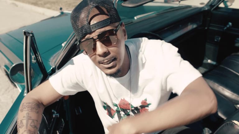 Man in white t-shirt and sunglasses leaning against a teal vintage convertible car.