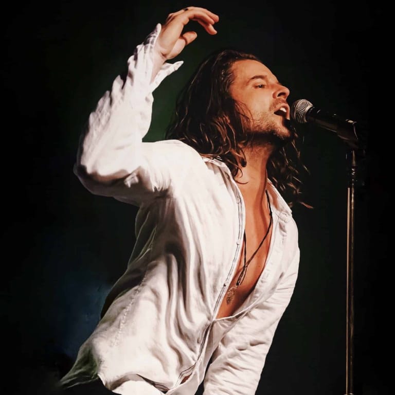 Male performer in white shirt singing at microphone with dramatic arm gesture against dark background.