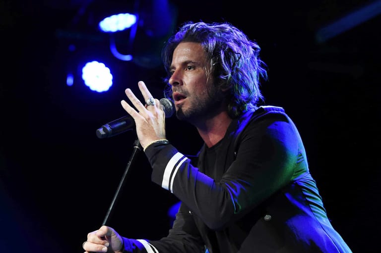 Male performer singing into microphone under blue stage lighting, gesturing expressively.