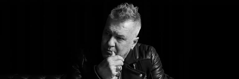 Man in black leather jacket with hand near mouth against dark background.