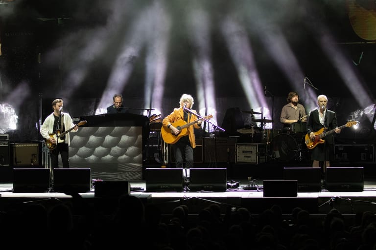 Five musicians perform on stage with dramatic purple and white spotlights beaming overhead.