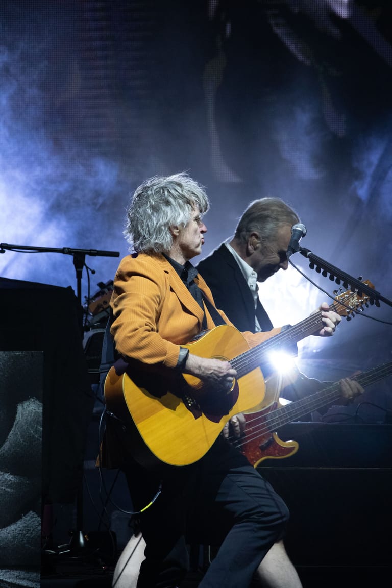 Two musicians perform on stage, one in a yellow jacket playing acoustic guitar, the other at a keyboard, bathed in blue stage lighting.