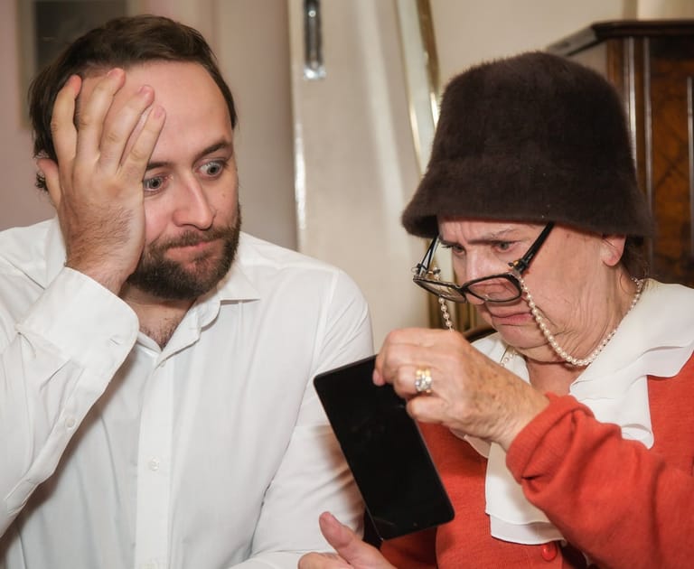 Two people indoors examine a small dark object together, one wearing a white shirt and the other a black hat and red garment.
