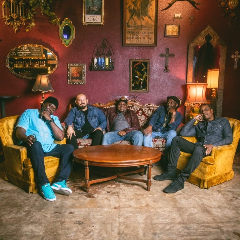 Seven people seated on yellow armchairs in a colourful bohemian lounge with ornate wall decorations and warm lighting.