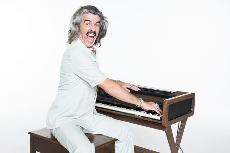 Person with grey hair in white suit playing vintage keyboard against white background.