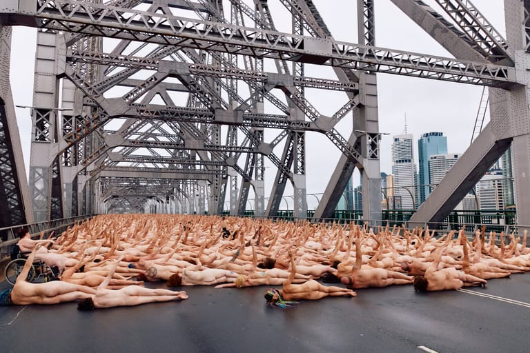 Large group of nude figures lying on ground beneath industrial bridge structure with city skyline.