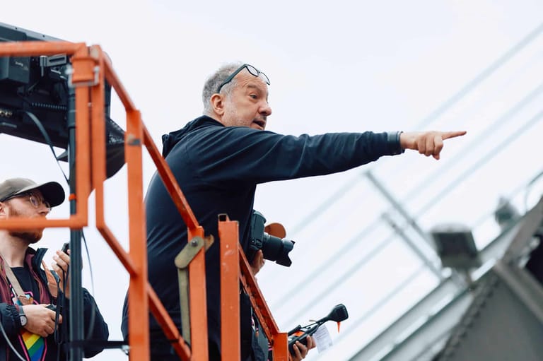 Man in dark jacket points outward from elevated orange platform at outdoor event.