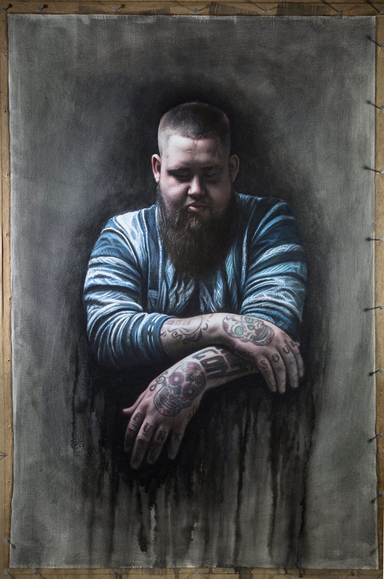 Bearded man with tattooed arms in striped shirt posed against dark, textured painted background.