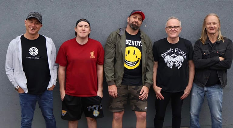 Five men in casual clothing stand in a line against a grey wall.