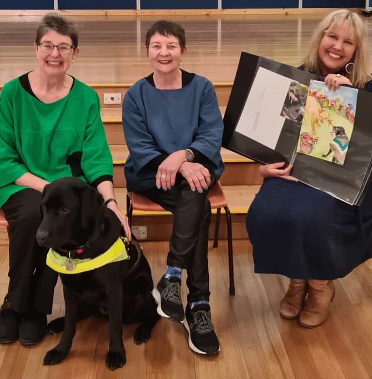 Three people sit on wooden bleachers with a black Labrador wearing a yellow service vest, displaying artwork and a framed photograph.